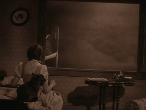 The Wizard of Oz - Dorothy Judy Garland looking at old woman knitting in rocking chair with cat in lap outside window in tornado animated gif
