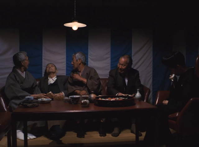 The Funeral - orange and white cat Kichi on lap of sleeping elderly man sitting at table with other people